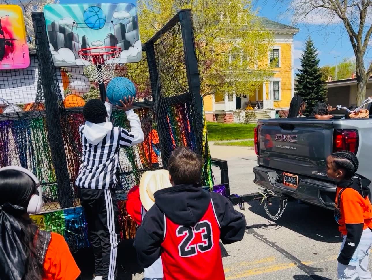 The hoops trailer hitched to a pickup truck at a parade while a child shoots at the city-skyline backboard.