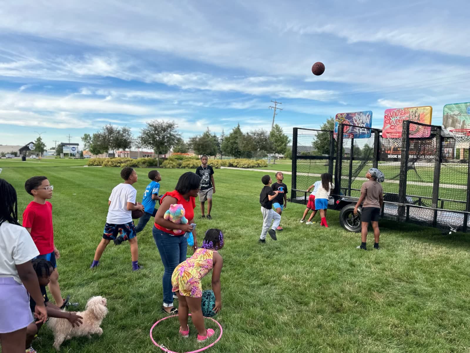 A dozen children playing at once around the hoops trailer on an open grass field at a community event.