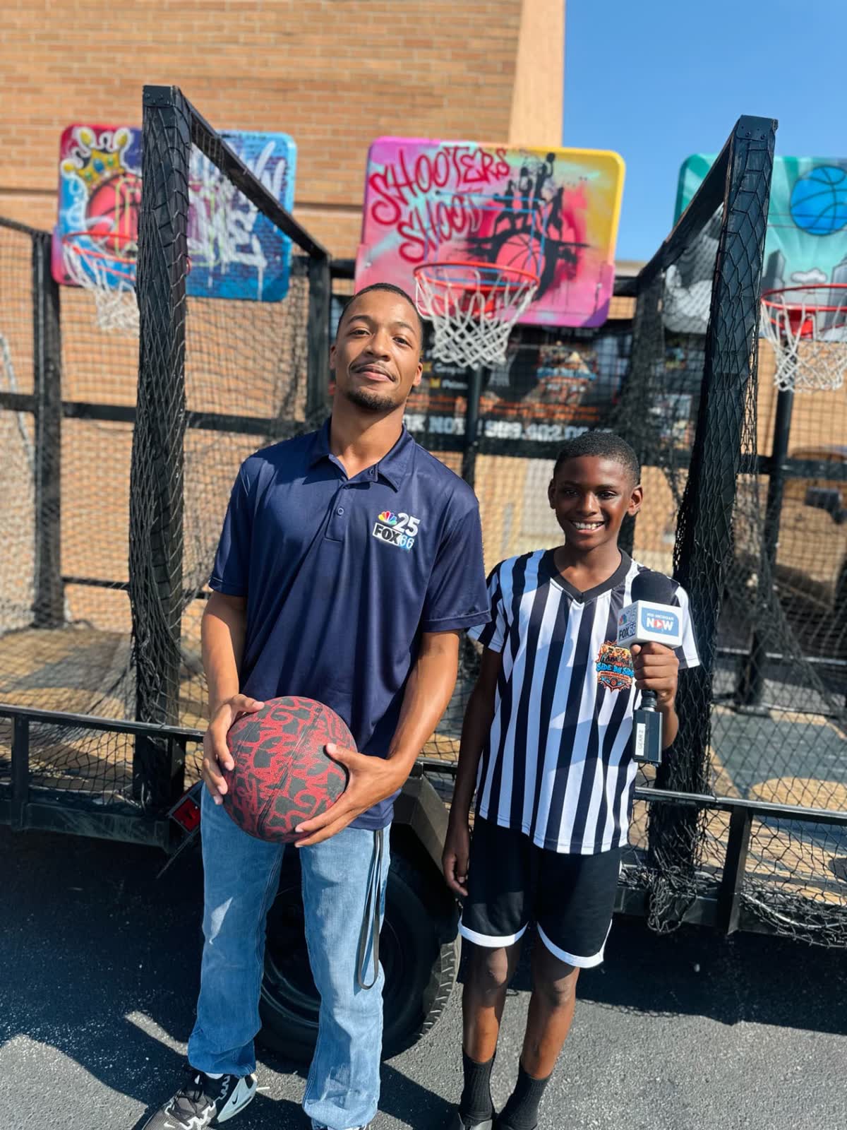 A local news reporter stands with a young Side By Side Mobile Hoops team member holding a station microphone in front of the trailer.